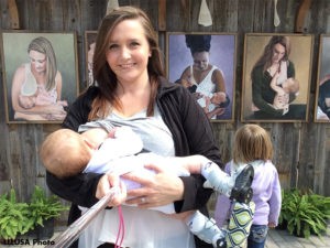Heather nursing her son, standing in front of photographs on a wall of nursing mothers. Heather's son is wearing his Boots and Bar.